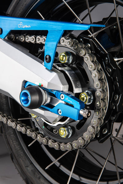 LighTech WAPHO406COB cobalt-blue rear axle slider installed, chain-side view; paddock stand, stand supports, chain adjusters, chain guard, coloured fasteners and motorcycle accessories are not included.