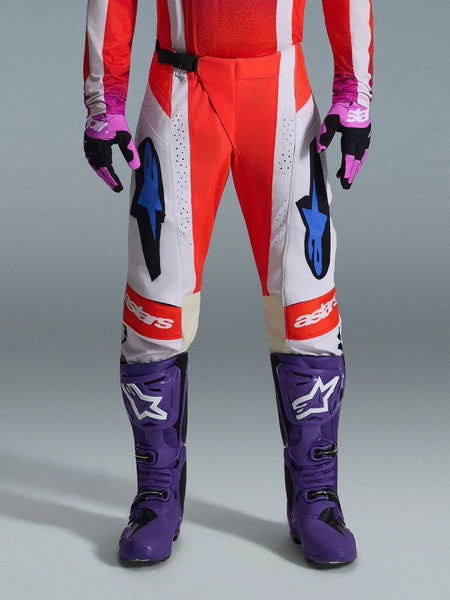 Alpinestars 2026 Techstar Knif Pants, Motocross Pants in Orange Fluo and Light Gray with Black accents, shown with purple motocross boots and gloves, featuring blue star logos and multi-panel construction for off-road performance