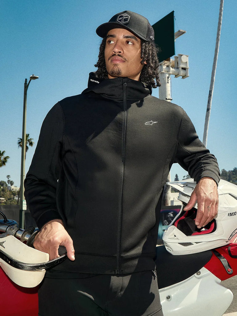 Alpinestars Cypher Hoodie, black zip-up lifestyle fleece, high-neck hood with branding, small white Astars logo on chest, worn with a black cap by a rider holding a white motocross helmet, casual motorsport streetwear