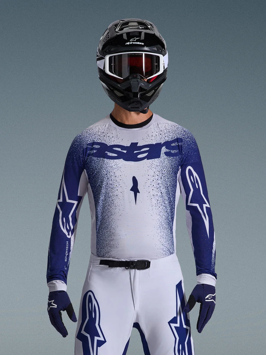 Motocross rider wearing the Alpinestars 2026 Supertech Scenz Jersey, Light Gray and Dark Blue with a speckled gradient design and bold 'astars' branding. Worn with matching white motocross pants, dark blue gloves, and a black helmet with goggles, coordinated outfit for professional off-road racing.