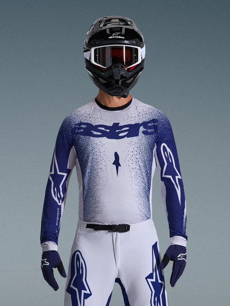Motocross rider wearing the Alpinestars 2026 Supertech Scenz Jersey, Light Gray and Dark Blue with a speckled gradient design and bold 'astars' branding. Worn with matching white motocross pants, dark blue gloves, and a black helmet with goggles, coordinated outfit for professional off-road racing.