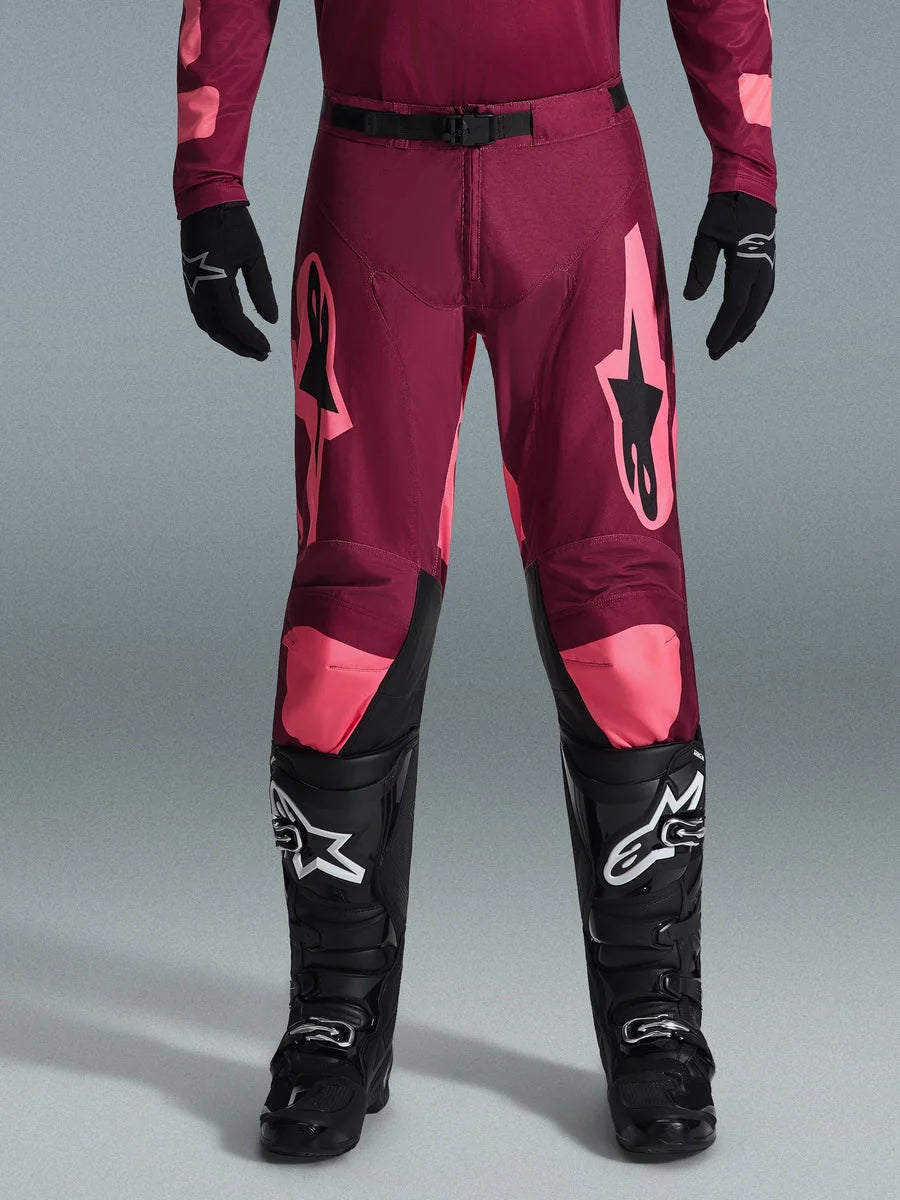 Alpinestars 2026 Racer Riway Pants, Motocross Pants, Dark Red Coral, worn with black gloves and boots, featuring large coral Alpinestars logos on thighs, black waist buckle, and pre-curved leg design for off-road racing