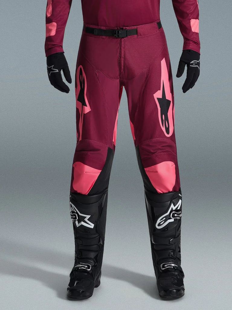 Alpinestars 2026 Racer Riway Pants, Motocross Pants, Dark Red Coral, worn with black gloves and boots, featuring large coral Alpinestars logos on thighs, black waist buckle, and pre-curved leg design for off-road racing
