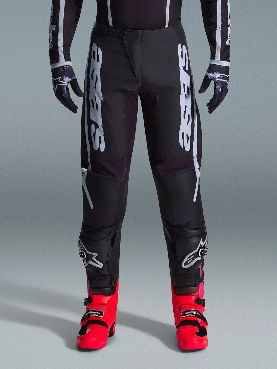 Alpinestars 2026 Fluid Apex Pants, Black Motocross Pants with large White Lettering, worn with black knee protection and vibrant red boots, lightweight 600D polyfabric chassis, sport-fit design for off-road racing
