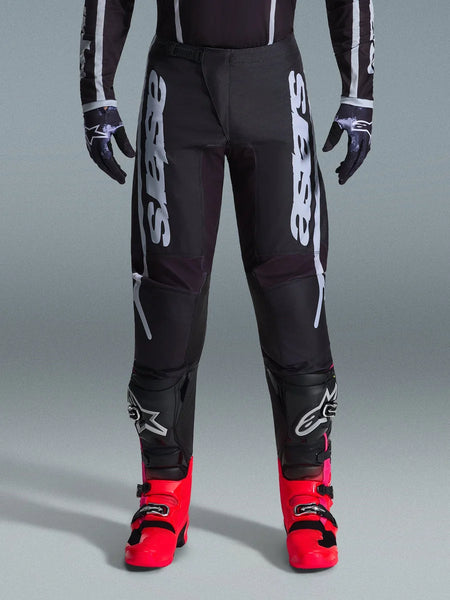 Alpinestars 2026 Fluid Apex Pants, Black Motocross Pants with large White Lettering, worn with black knee protection and vibrant red boots, lightweight 600D polyfabric chassis, sport-fit design for off-road racing