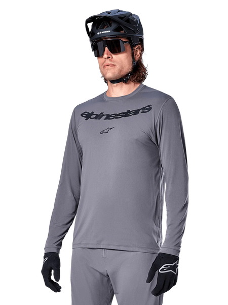 Alpinestars A-Dura Rocker Long Sleeve Jersey, Gray MTB Mountain Biking Jersey, worn with black helmet and gloves, featuring black wordmark on the chest, sporty cycling apparel for off-road riding, plain background