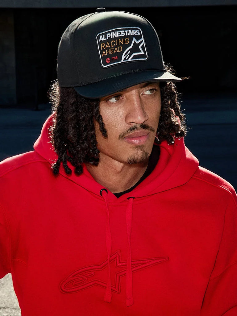Man wearing Alpinestars Intuitive Snapback Hat in Black, featuring a structured crown and 'Racing Ahead' logo embroidery patch, paired with a bright red hoodie, casual lifestyle streetwear