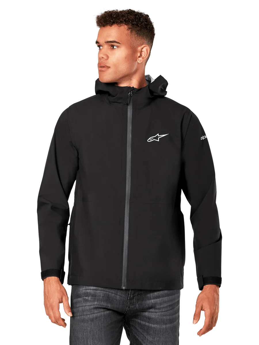Male model wearing Alpinestars Kascade Rain Jacket, Black, white chest logo and sleeve branding, hooded rain shell with coated front zipper and velcro cuffs, paired with dark jeans, casual motorsport-inspired streetwear
