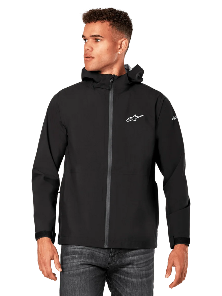 Male model wearing Alpinestars Kascade Rain Jacket, Black, white chest logo and sleeve branding, hooded rain shell with coated front zipper and velcro cuffs, paired with dark jeans, casual motorsport-inspired streetwear