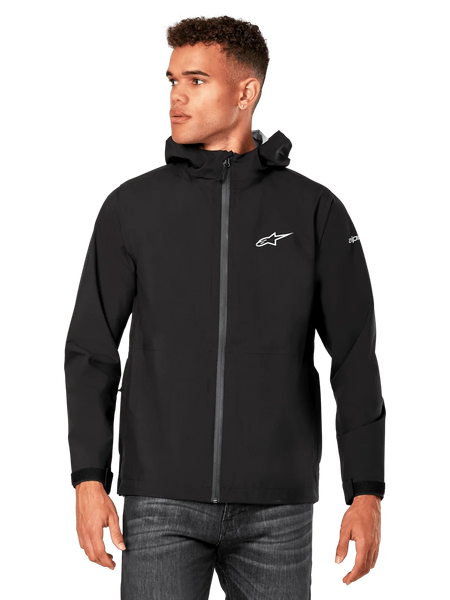 Male model wearing Alpinestars Kascade Rain Jacket, Black, white chest logo and sleeve branding, hooded rain shell with coated front zipper and velcro cuffs, paired with dark jeans, casual motorsport-inspired streetwear