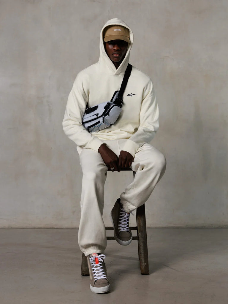 Alpinestars Essentials Ageless Hoodie, Off-White, worn by a model sitting on a stool with matching fleece joggers, tan ASTARS cap, and cross-body bag, casual motorsport-inspired streetwear