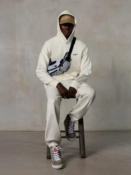 Alpinestars Essentials Ageless Hoodie, Off-White, worn by a model sitting on a stool with matching fleece joggers, tan ASTARS cap, and cross-body bag, casual motorsport-inspired streetwear