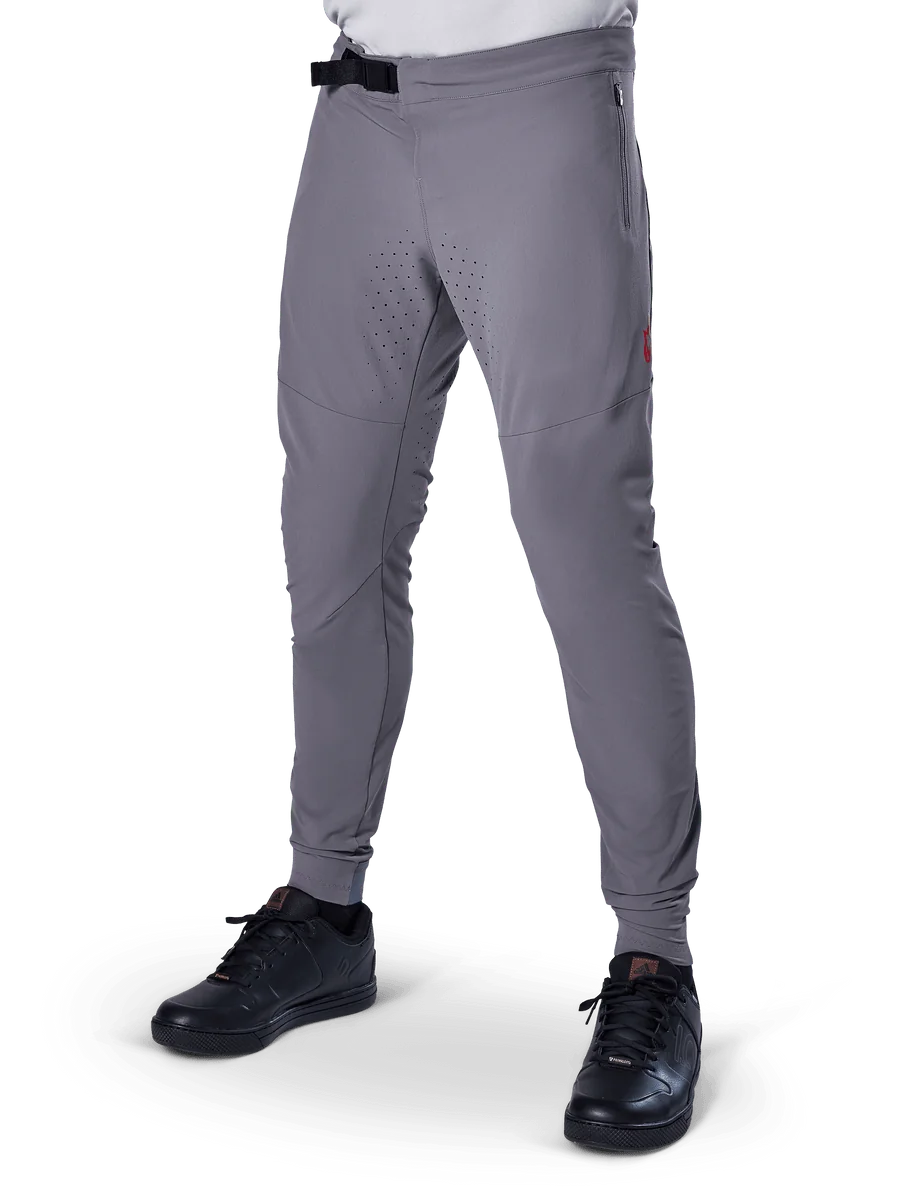 Alpinestars A-Aria Elite Pants, Dark Gray MTB Pants, laser perforated panels on thighs, black magnetic buckle closure, zippered pocket, red logo detail, slim performance fit, worn with black shoes