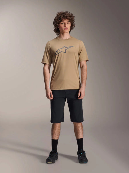 Model wearing Alpinestars A-Dura SS Jersey in tan and A-Dura Shorts in black, featuring a grey star logo on the chest, paired with black socks and mountain biking shoes, complete MTB riding apparel in a studio setting.