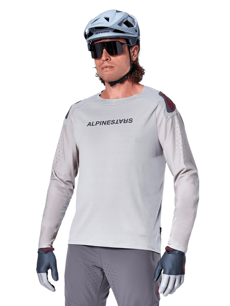 Alpinestars A-Aria Polartec Switch Jersey Long Sleeve, Light Gray cycling jersey with black chest logo and red shoulder accent, laser-perforated sleeves for airflow, worn with a light blue helmet, sunglasses, and gray pants, performance mountain bike apparel for trail riding
