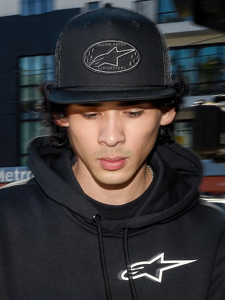 Alpinestars Contender Trucker Hat, black lifestyle cap with poly mesh back, structured crown featuring an embroidered Astars logo patch, worn by a man with a black Alpinestars hoodie, casual motorsport-inspired streetwear.