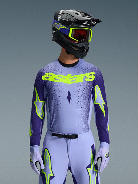 Alpinestars 2026 Supertech Scenz Jersey, Motocross Jersey, Purple and Yellow Fluo, worn by a rider with a matching black and purple helmet, neon yellow goggles, and purple racing pants, featuring neon yellow astars branding on the chest and star logos on the sleeves.