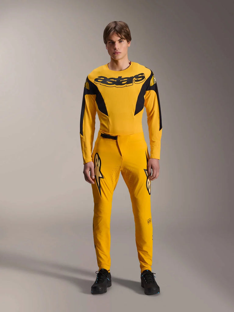 Limited Edition A-Supra Melt Gold Gear Set, Alpinestars Mountain Bike Apparel, Dark Yellow and Black, Model wearing long-sleeve jersey with bold 'astars' chest logo and matching performance pants, featuring laser-cut ventilation on inner thighs and a secure ratchet waist closure, professional MTB racing kit