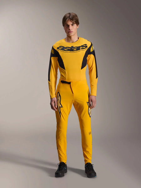 Limited Edition A-Supra Melt Gold Gear Set, Alpinestars Mountain Bike Apparel, Dark Yellow and Black, Model wearing long-sleeve jersey with bold 'astars' chest logo and matching performance pants, featuring laser-cut ventilation on inner thighs and a secure ratchet waist closure, professional MTB racing kit