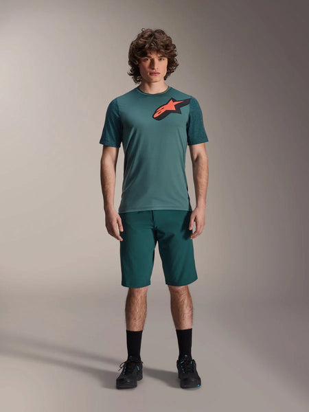 Alpinestars A-Dura Shorts in Dark Pine, men's mountain bike shorts, shown on a model with a matching teal short-sleeve jersey featuring a red and black star logo, black socks and cycling shoes, casual sporty mountain biking apparel