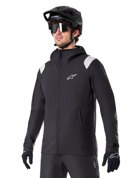 Alpinestars A-Dura Rain Jacket, Mountain Bike Rain Jacket, Black with light gray shoulder accents, worn by a rider with a black MTB helmet and sunglasses, white chest logo, waterproof construction, coordinated with black pants and gloves for off-road cycling