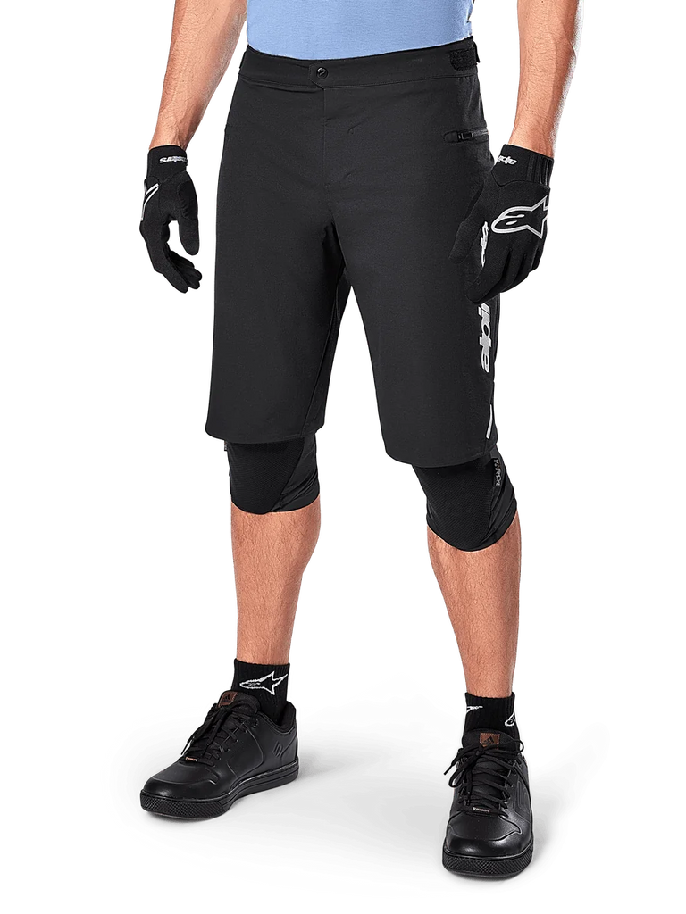 Alpinestars A-Dura Elite Shorts, Black MTB Mountain Biking Shorts, worn with light blue shirt and black knee protection, featuring a secure snap closure and zippered pocket, paired with black cycling shoes and gloves for a complete off-road outfit.